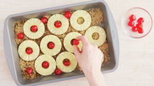 Baking pan with brown sugar and sliced pineapple with a hand putting a cherry in center of each slice of pineapple prior to baking the yellow cake